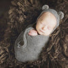 A newborn baby is sleeping inside a knitted gray bear hat and wrap set, nestled in dark brown fur.