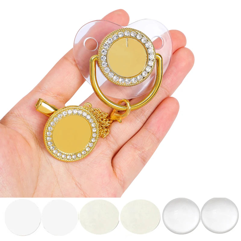 A hand holds a gold-toned baby pacifier and clip set with rhinestone accents.