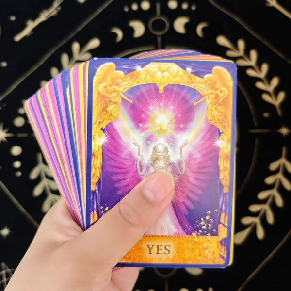 A hand holds a tarot card from an oracle deck featuring a golden angel motif and the word YES on a purple background.