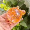 A hand holds a translucent orange squishy star turtle fidget ball with sparkling details against a blurred green background.