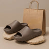 A pair of taupe EVA platform slides resting on light rocks next to a brown paper shopping bag.