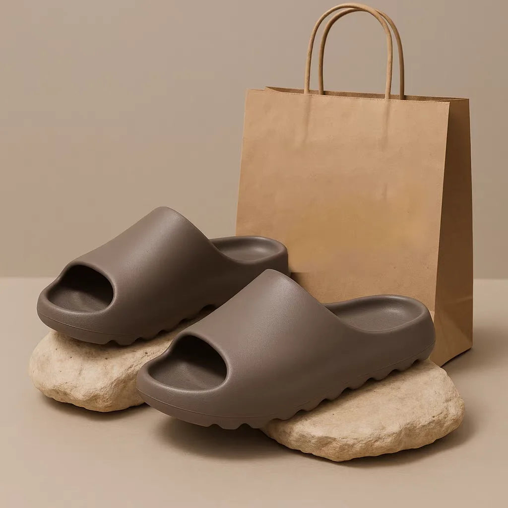 A pair of taupe EVA platform slides resting on light rocks next to a brown paper shopping bag.
