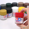 A hand holds a bottle of Tamiya Color Flat Red acrylic paint for model kits, surrounded by various paint pots.