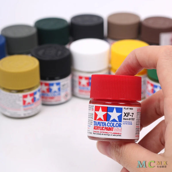 Mano sostiene una botella de pintura acrílica Tamiya Color Flat Red para maquetas, rodeada de varios botes de pintura.
