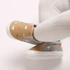 A close-up of a baby first walker sneaker with a tan upper featuring small dinosaur prints and a white sole.
