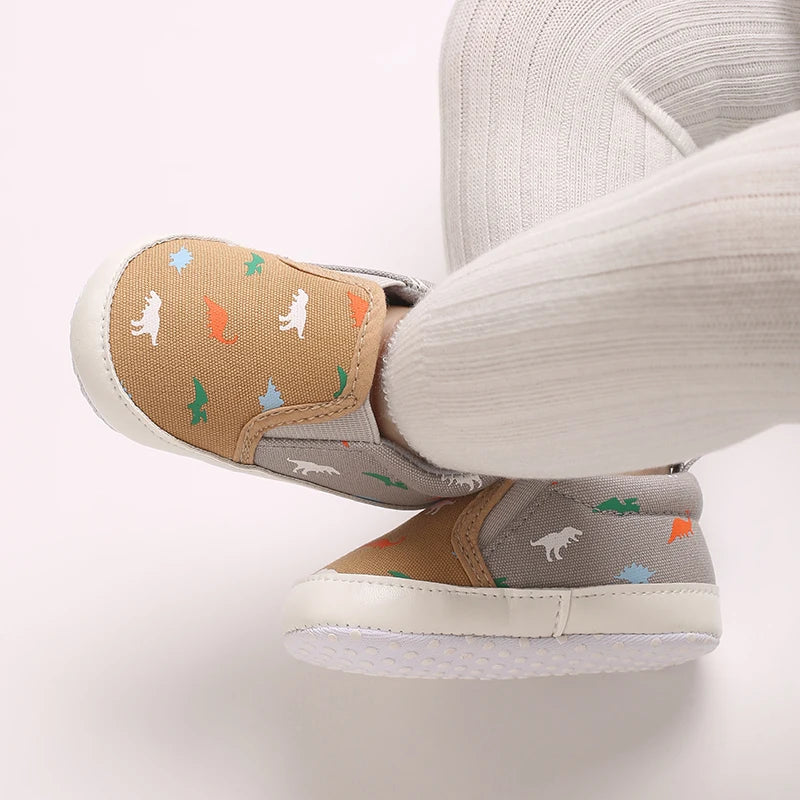 A close-up of a baby first walker sneaker with a tan upper featuring small dinosaur prints and a white sole.