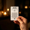 A hand holds a tarot-style oracle card featuring celestial symbols and text against a dark background.