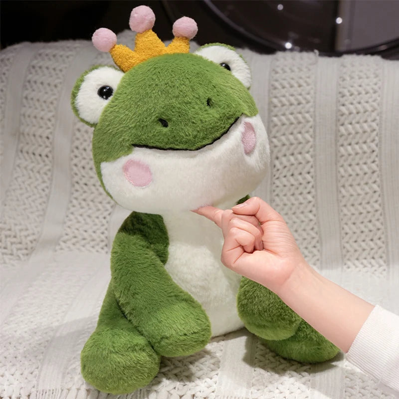 A hand gently touches a cute green frog prince plush toy with a yellow crown.