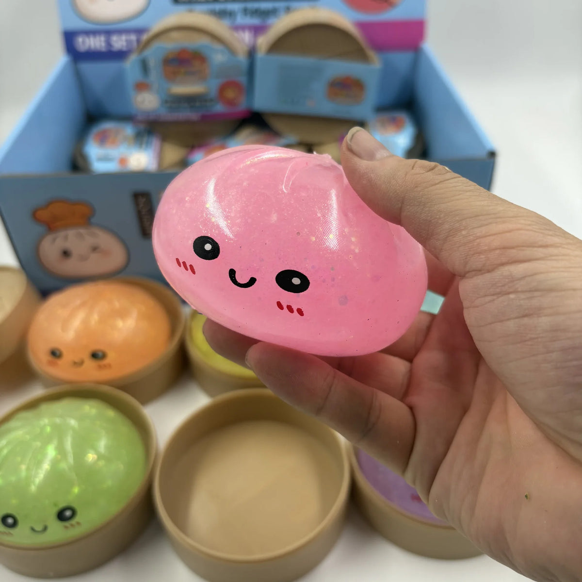 A hand holds a pink squishy dumpling toy with simple facial features, surrounded by other soft sensory toys.