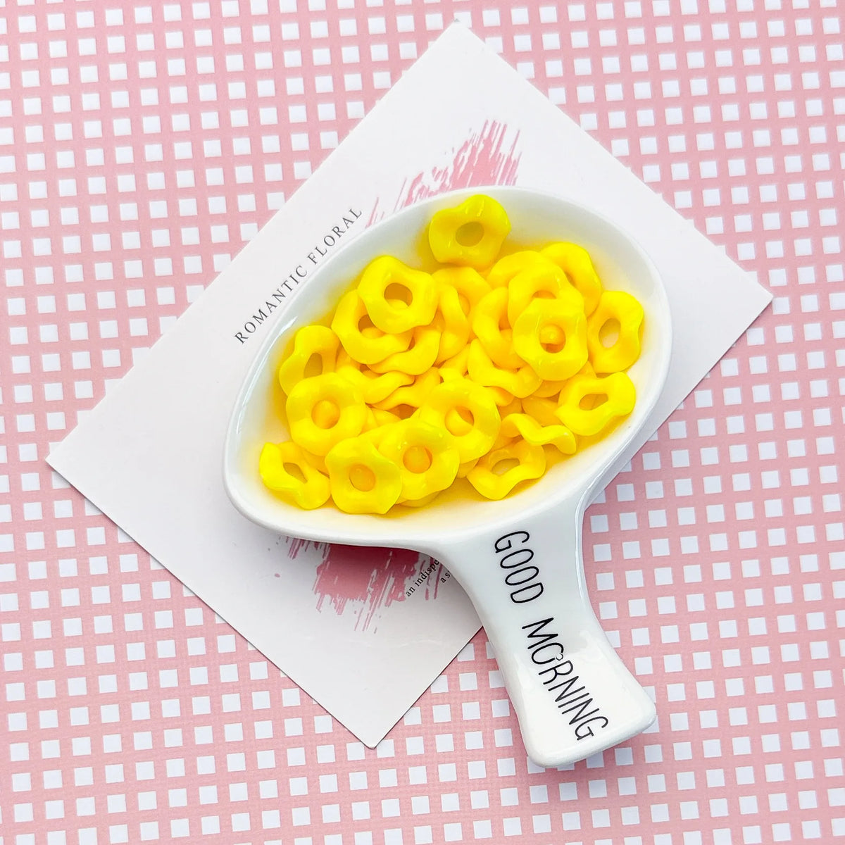 A white spoon holds bright yellow acrylic beads with a floral edge on a pink patterned background.