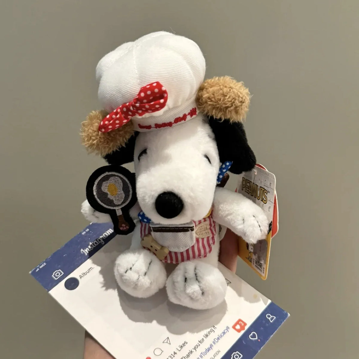 A white Snoopy plush charm with a red bow and brown accents is displayed on a white card.