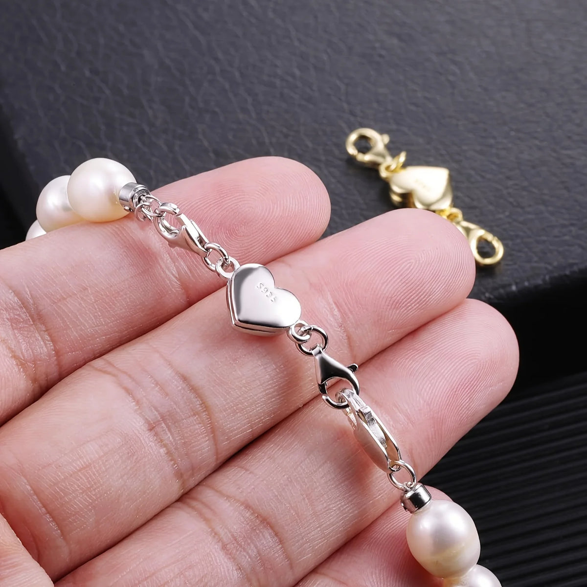 A hand holds a silver heart-shaped magnetic clasp necklace with pearl beads and a gold jump ring.
