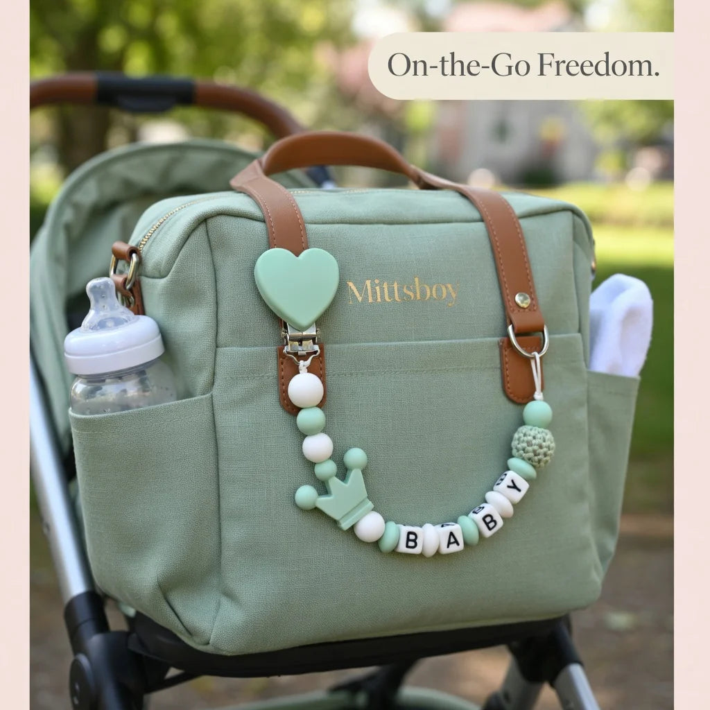A light green diaper bag featuring a personalized silicone pacifier clip with a baby name charm.