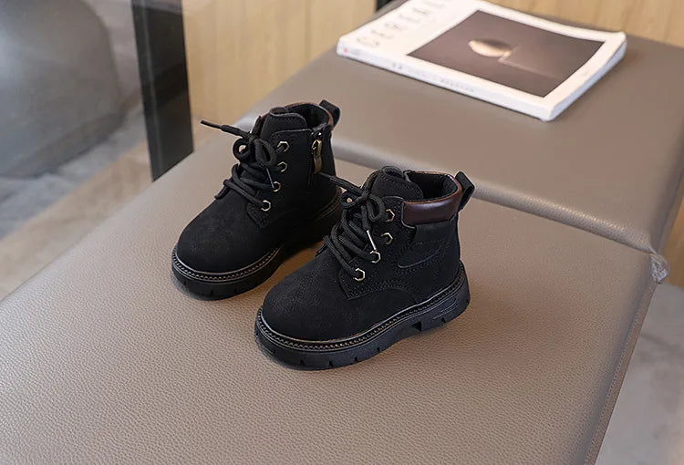 A pair of black kids' winter leather boots with laces and a brown cuff resting on a gray surface.