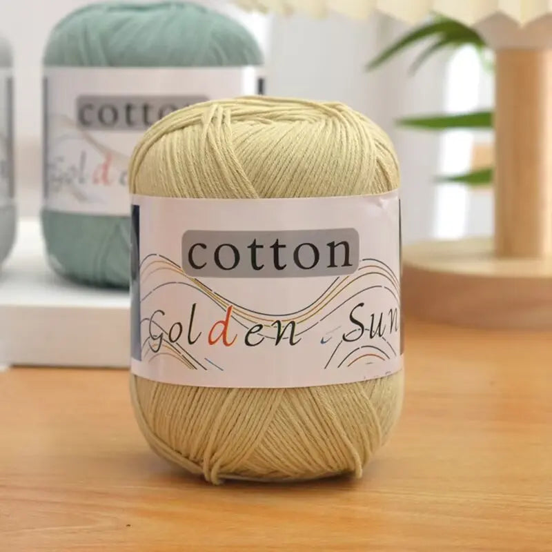 A ball of light beige cotton knitting yarn labeled Golden Sun is displayed on a wooden surface.