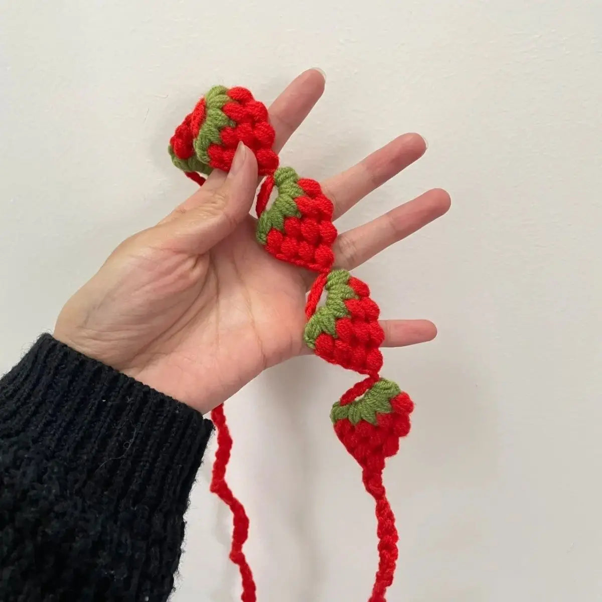 A hand is holding a red strawberry crochet headband with green leaves and a wide elastic band.