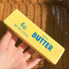 A yellow butter stick with the text 4OZ NET WT 113 G and BUTTER printed on it, held by a hand against a woven background.