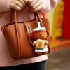A brown leather handbag with a teddy bear plush keychain charm is held by a person wearing a pink sweater.