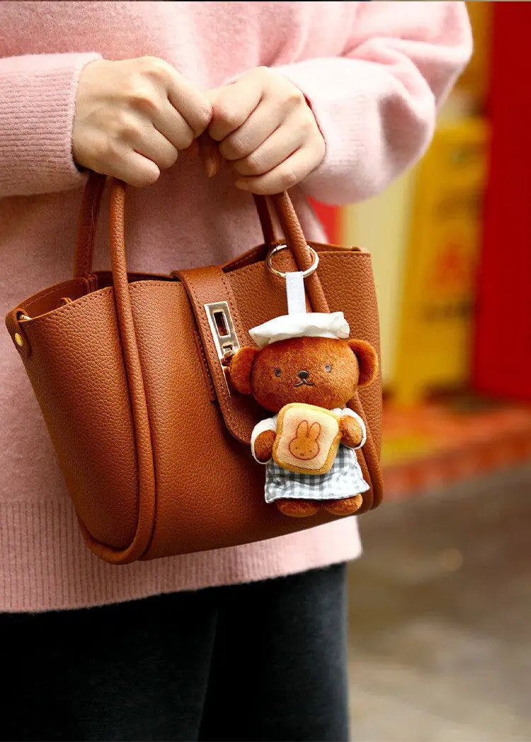 A brown leather handbag with a teddy bear plush keychain charm is held by a person wearing a pink sweater.