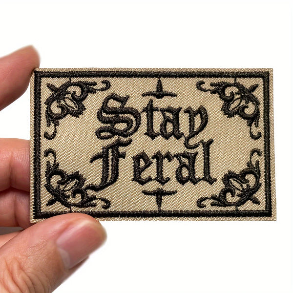 Ein bestickter Bügelaufnäher mit dem schwarzen Schriftzug „Stay Feral“ auf einem beigen Stoffhintergrund