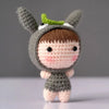 A handmade crochet doll kit featuring a gray body, brown hair, and green ears with a white face patch.