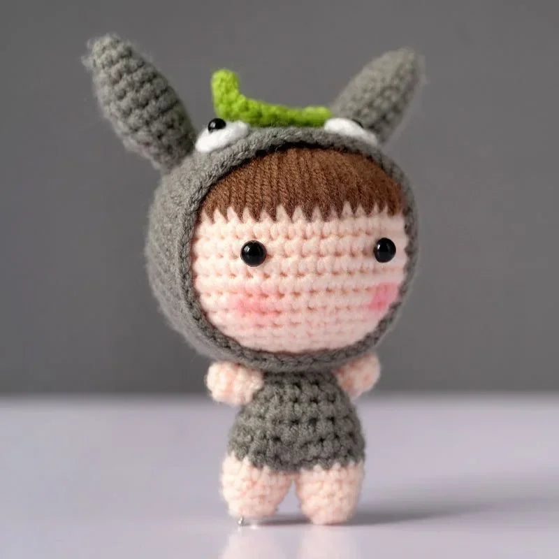 A handmade crochet doll kit featuring a gray body, brown hair, and green ears with a white face patch.