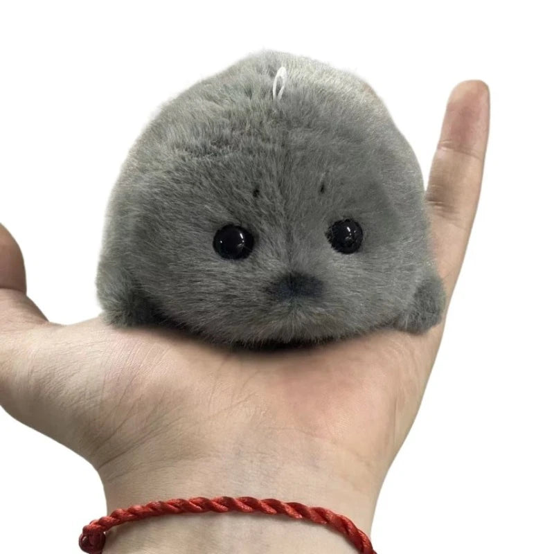 A small, round grey seal plush toy is held in a person's hand.