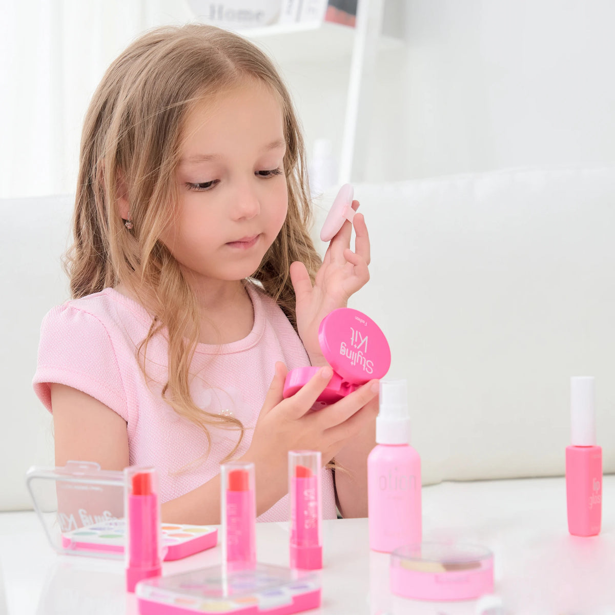 A young girl is holding a pink kids pretend makeup kit with various cosmetic items on a white table.