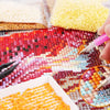 A hand is arranging colorful AB rhinestones on a diamond painting canvas, showing various shades of pink, red, and yellow.