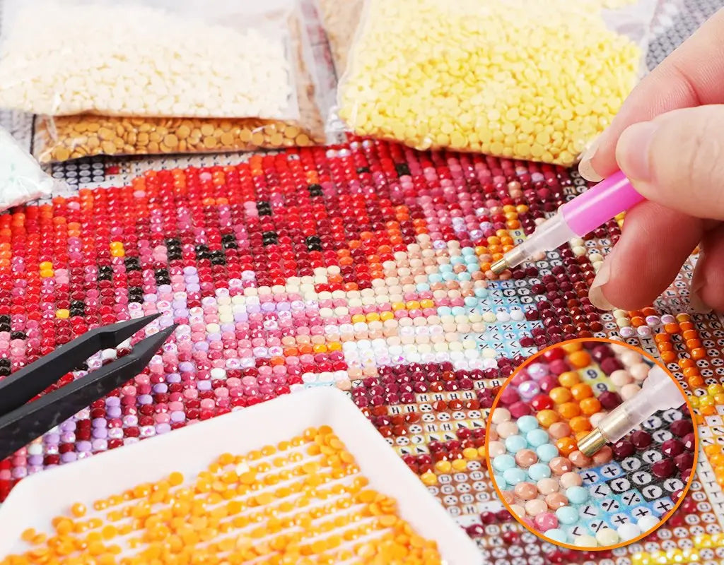 A hand is arranging colorful AB rhinestones on a diamond painting canvas, showing various shades of pink, red, and yellow.