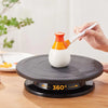 A hand is rotating a white and orange ceramic cake decorating tool on a black turntable for food preparation.