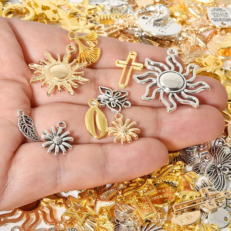 A hand holds various mixed metal charms including suns, crosses, and floral shapes for DIY jewelry making