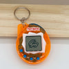 An orange interactive 3D virtual pet toy with a digital screen and keyring is displayed on a wooden surface.