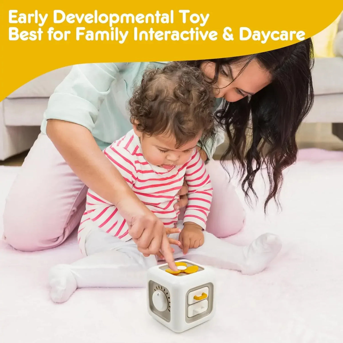 A baby is interacting with a white sensory cube toy on a pink blanket while being held by an adult.