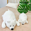 A white plush polar bear stuffed animal lying on a beige surface next to green foliage.