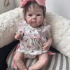 A lifelike 18-inch reborn baby doll with hand-painted details wearing a floral dress and pink bow, sitting on a white fluffy 