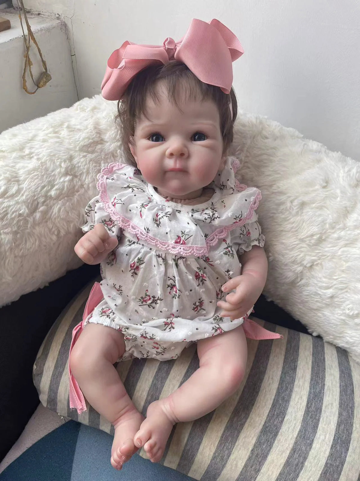 A lifelike 18-inch reborn baby doll with hand-painted details wearing a floral dress and pink bow, sitting on a white fluffy 