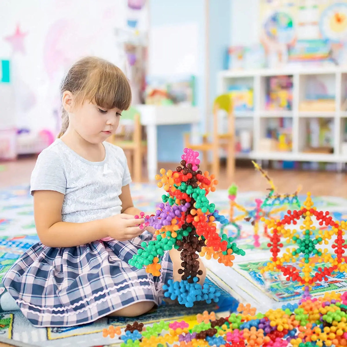 A young girl is building a colorful structure with plum blossom snowflake blocks on a mat in a classroom setting.