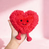A hand holds a bright red, fluffy heart-shaped plush cushion with pink feet against a light pink background.