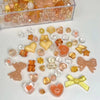 A collection of mixed acrylic bow and flower beads in various shades of orange, yellow, and clear are displayed on a white su