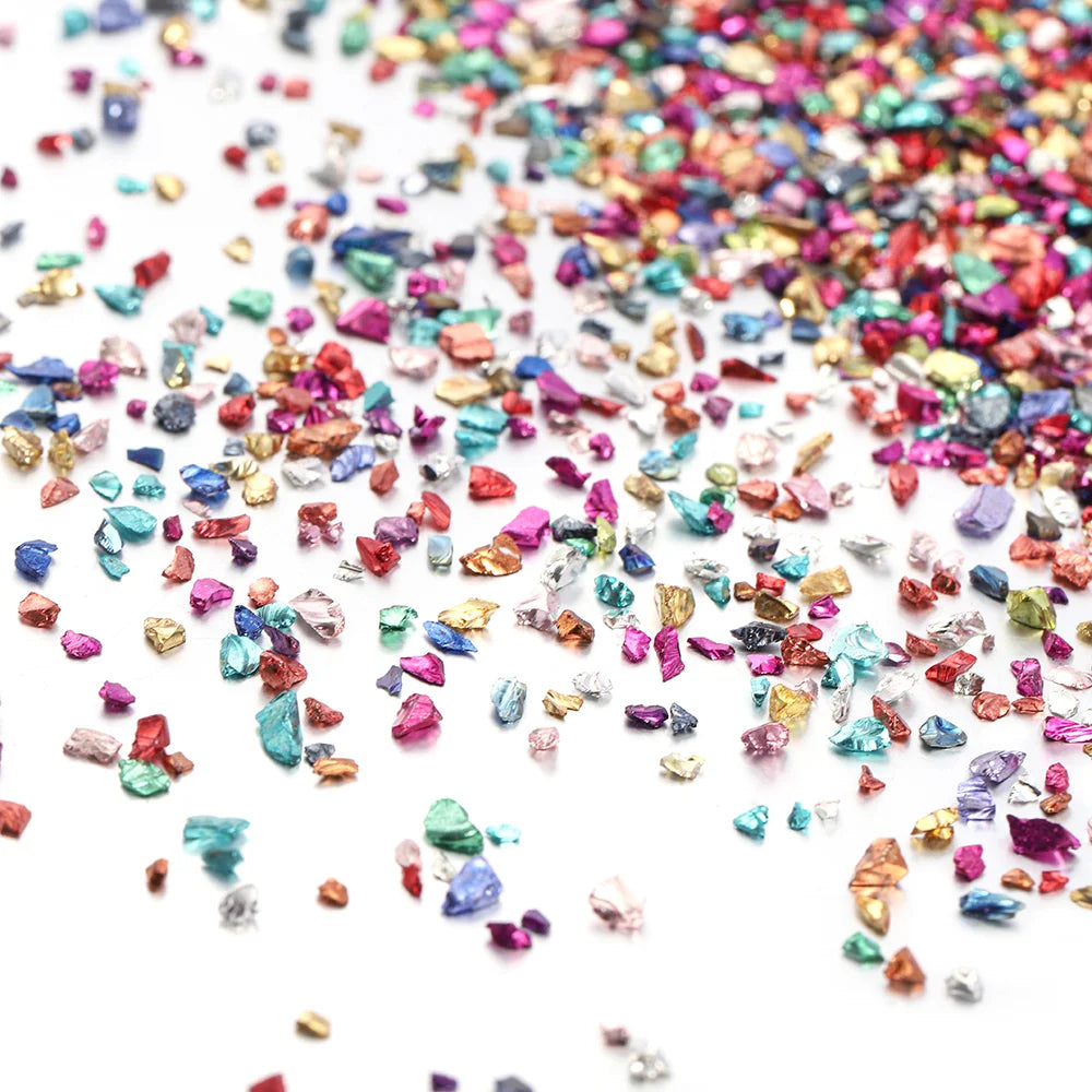 A close-up of multicolored crushed glass stones in various shades of pink, blue, and gold scattered on a white surface.