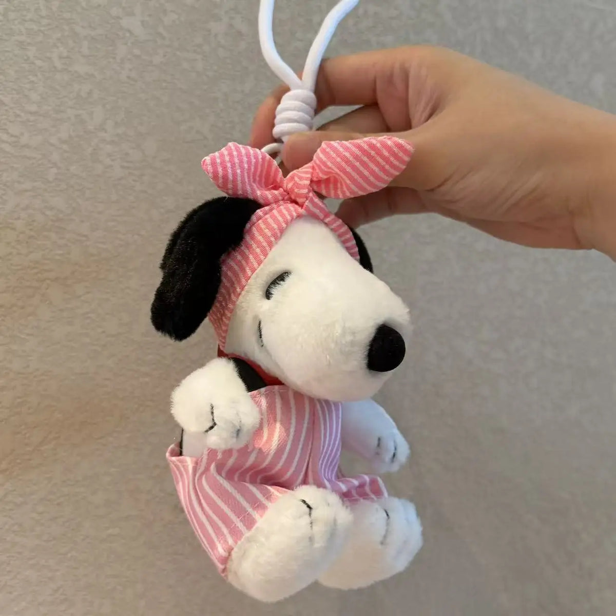 A white Snoopy plush toy with a pink and white striped bow and outfit is being held by a hand.