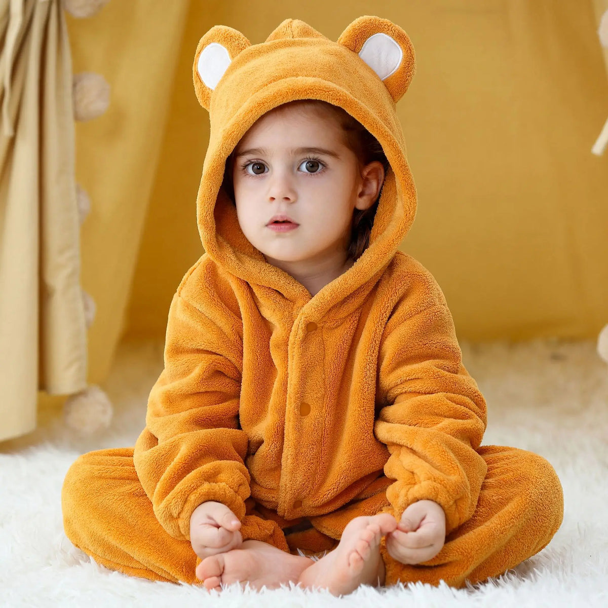 A baby wearing an adorable orange hooded romper resembling a fox, sitting on a white fluffy surface.
