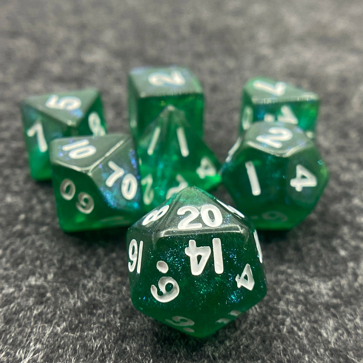 A collection of seven iridescent green acrylic polyhedral dice scattered on a dark gray surface.