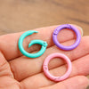 A hand holds several colorful metal O-ring spring clasps in various shades of turquoise, purple, and pink.