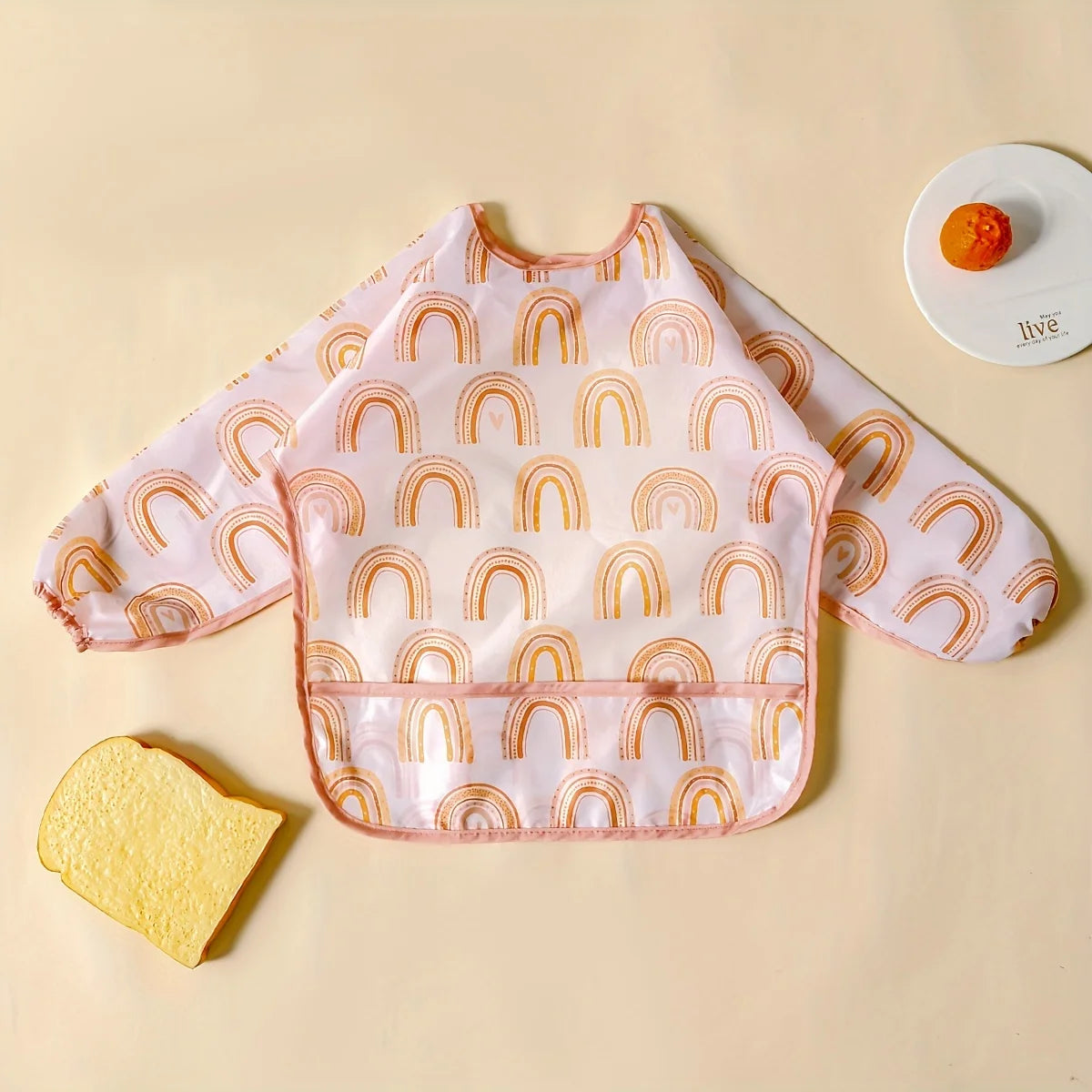 A pink waterproof baby bib with long sleeves featuring a rainbow pattern, placed next to a piece of yellow sponge cake.