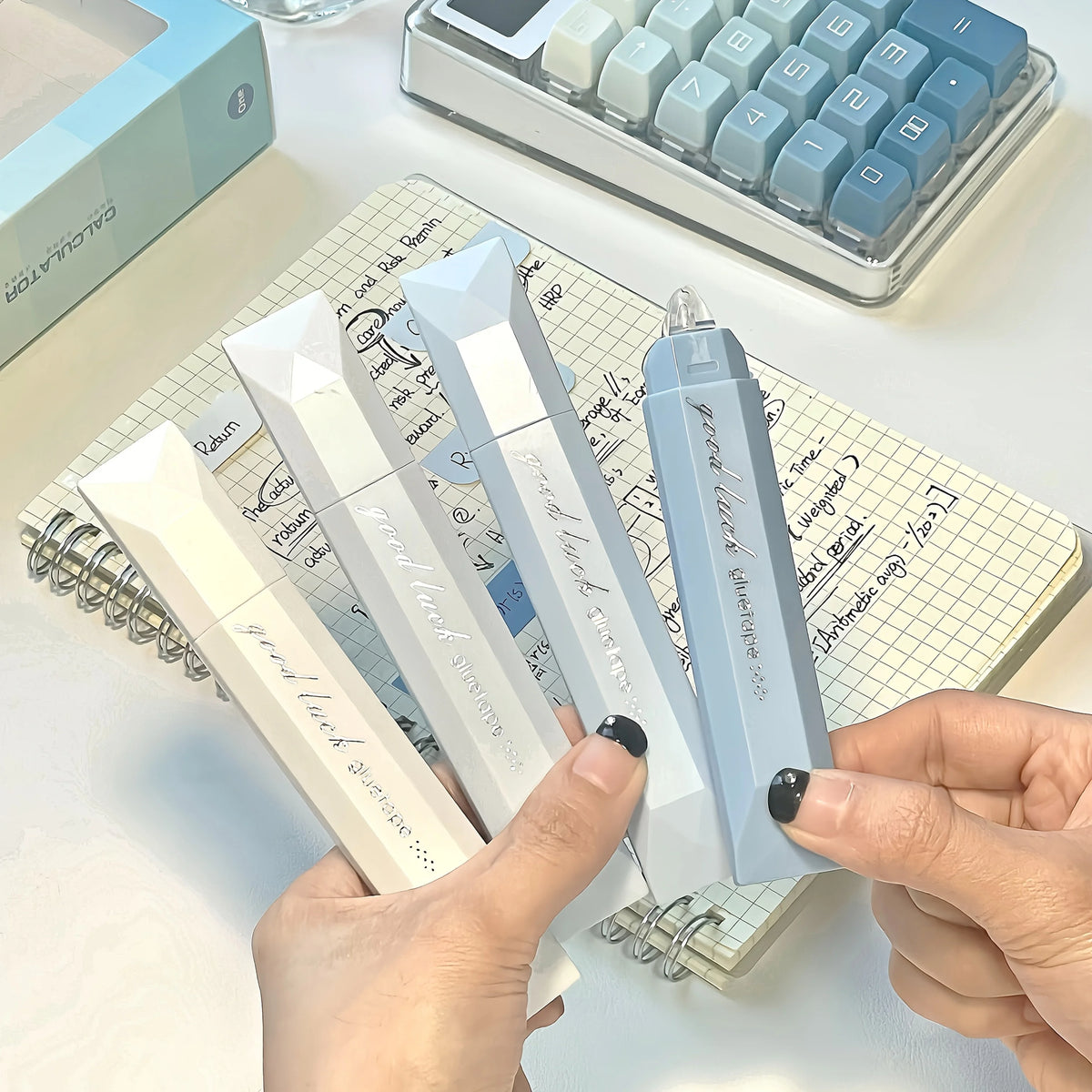 A hand holds a light blue adhesive dot tape glue roller next to a spiral notebook and a calculator.