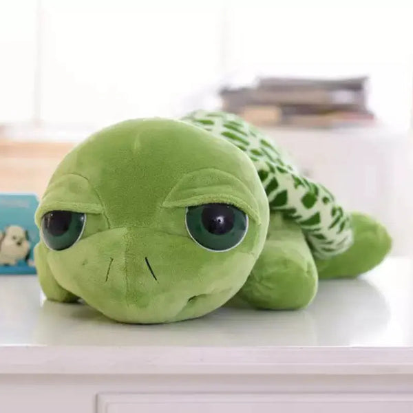 A soft green plush toy of a turtle with large eyes sitting on a white surface.