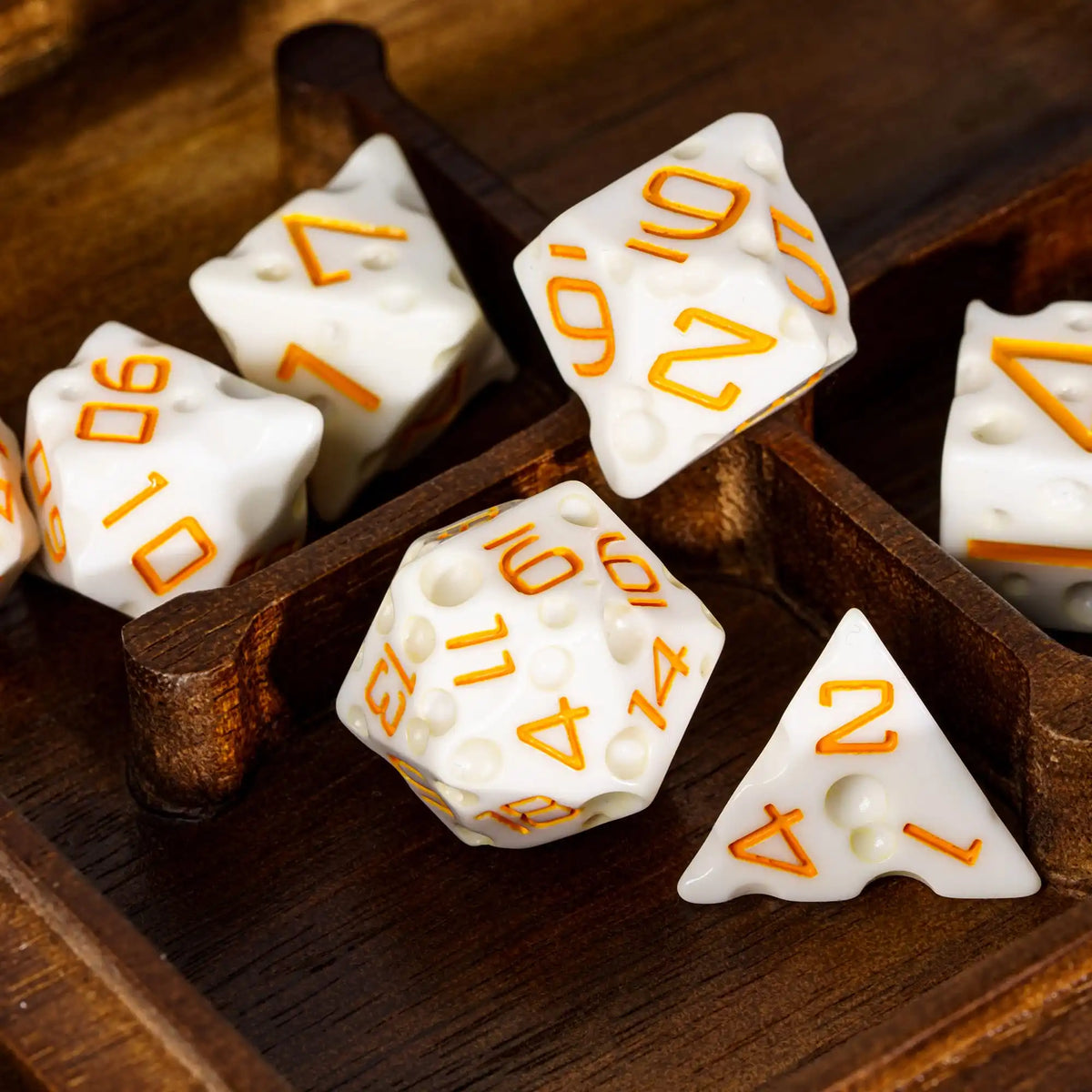 A set of seven white and orange polyhedral dice resting in a wooden box.