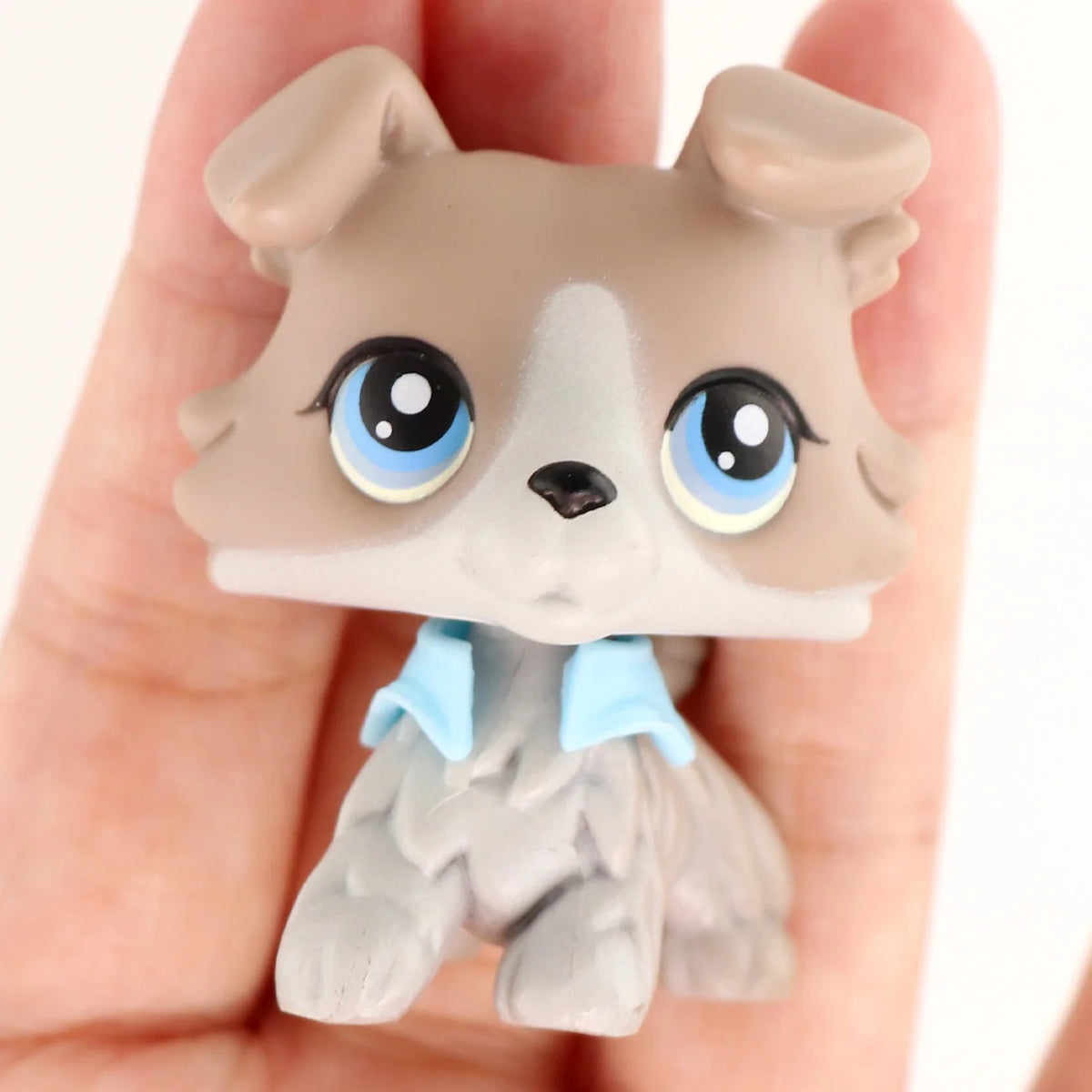 A small, light brown miniature pet animal figure with blue eyes and a light blue collar is held in a person's hand.